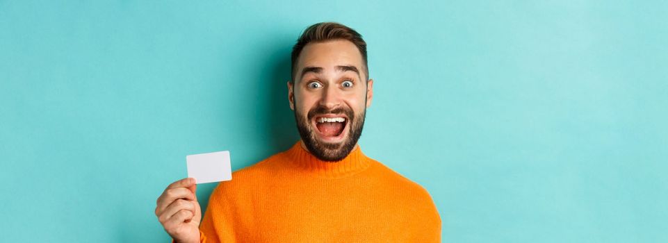 Close-up of excited caucasian man showing his credit card, smiling and staring amazed, standing in orange sweater against turquoise background.