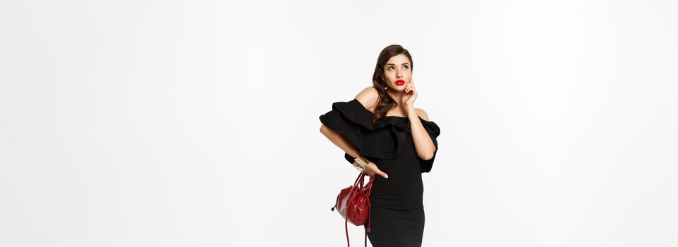 Beauty and fashion concept. Full length of beautiful and stylish woman in black dress thinking about shopping, looking at upper left corner, holding purse, white background.