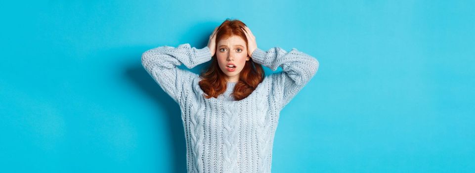 Nervous redhead girl standing overwhelmed, holding hands on head in panic and staring at camera, standing anxious against blue background.