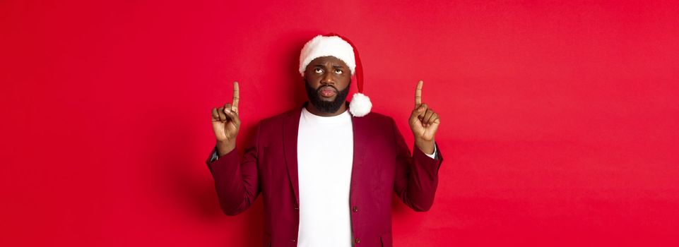 Christmas, party and holidays concept. Upset african-american guy in santa hat complaining, pointing fingers up and frowning disappointed, standing against red background.