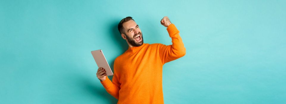 Cheerful winning man holding digital tablet, rejoicing and celebrating victory in game, making fist pump gesture, standing over light blue background.