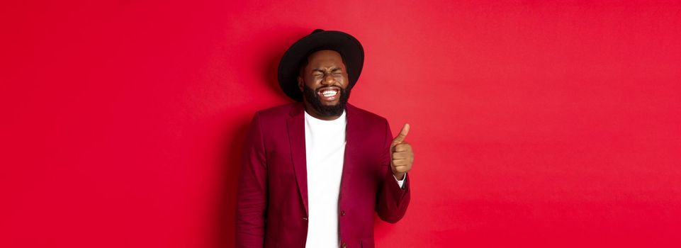 Christmas shopping and people concept. Handsome bearded Black man in party blazer showing thumb up, laughing and having fun, standing over red background.