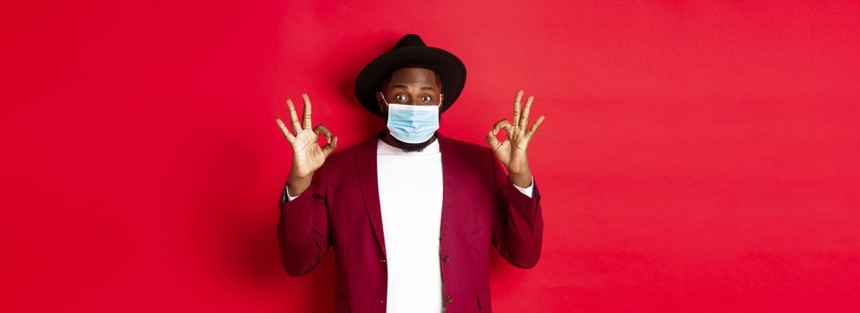 Covid-19 and fashion concept. Stylish african american man in hat and blazer, wearing face mask and showing okay sign, standing over red background.