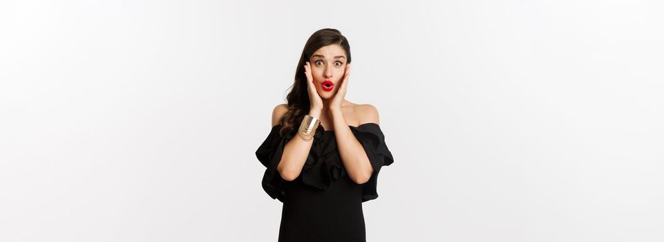 Beauty and fashion concept. Attractive woman in glamour black dress, jewelry and makeup, looking surprised and excited, standing over white background.