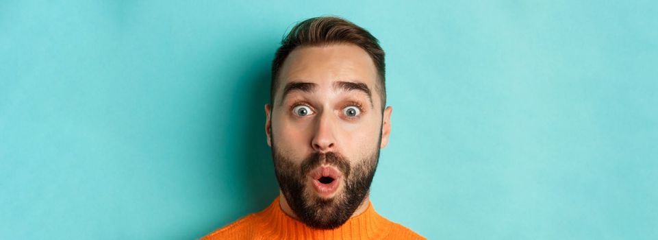 Headshot of handsome caucasian man with beard standing in orange sweater against turquoise background, saying wow and staring surprised at camera.