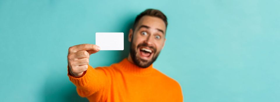 Close-up of handsome caucasian man going on shopping, showing credit card and smiling, standing over turquoise background.