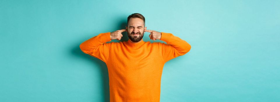 Displeased man shut ears and grimacing, loud neighbours, standing annoyed with noise, light blue background.