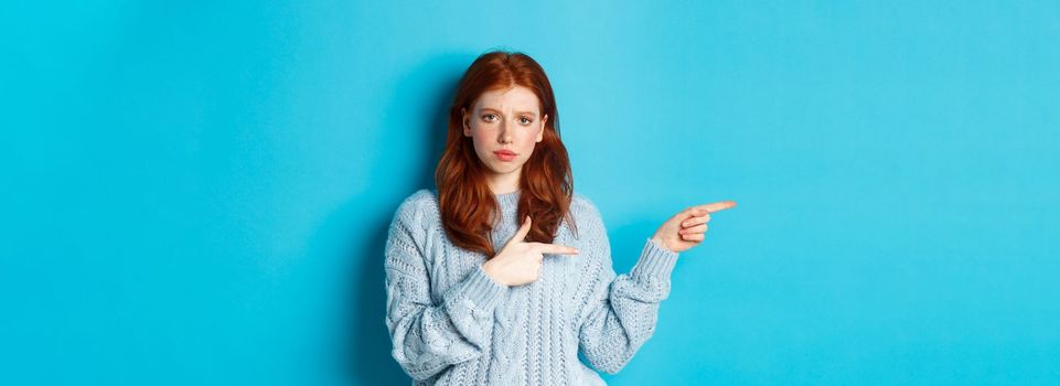 Skeptical and displeased redhead girl pointing fingers left with upset face, standing disappointed against blue background.