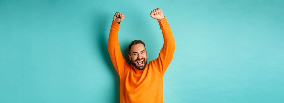 Waist-up shot of happy guy triumphing, raising hands up and rejoicing of winning, celebrating victory, standing against turquoise background.