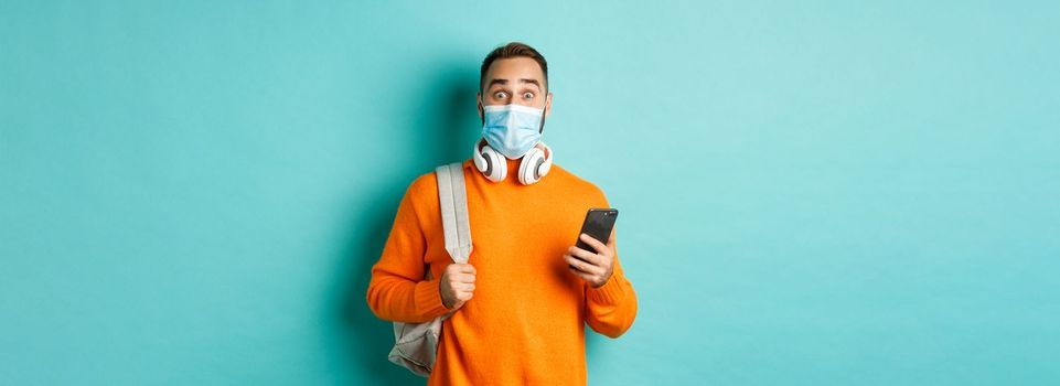 Young man in face mask using mobile phone, holding backpack, staring impressed at camera, standing against light blue background.