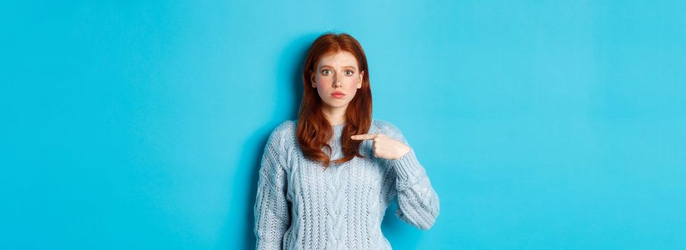 Nervous and confused redhead girl pointing at herself, standing in sweater against blue background.