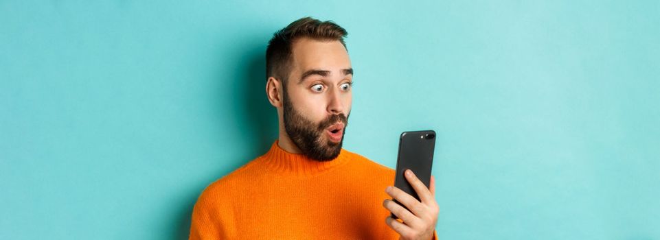 Close-up of caucasian man staring at phone screen with surprised face, wearing orange sweater, light blue background.