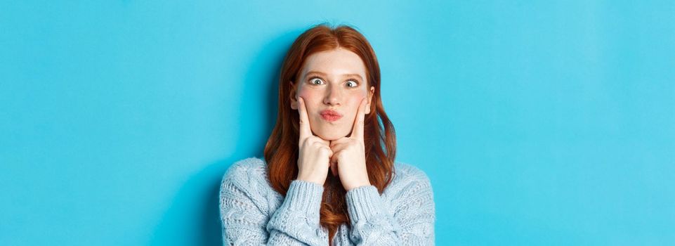 Close-up of funny redhead teen girl making faces, squinting and pocking cheeks, standing against blue background.