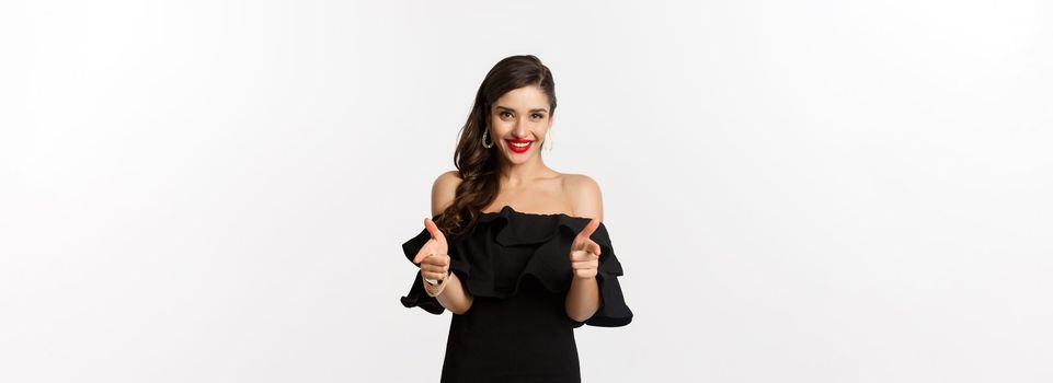 Fashion and beauty. Beautiful young woman in black dress, wearing makeup, pointing fingers at camera to congratulate or praise, smiling pleased, standing over white background.