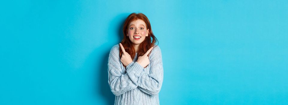 Excited redhead girl pointing fingers sideways, showing two choices and looking tempted at camera, standing against blue background.