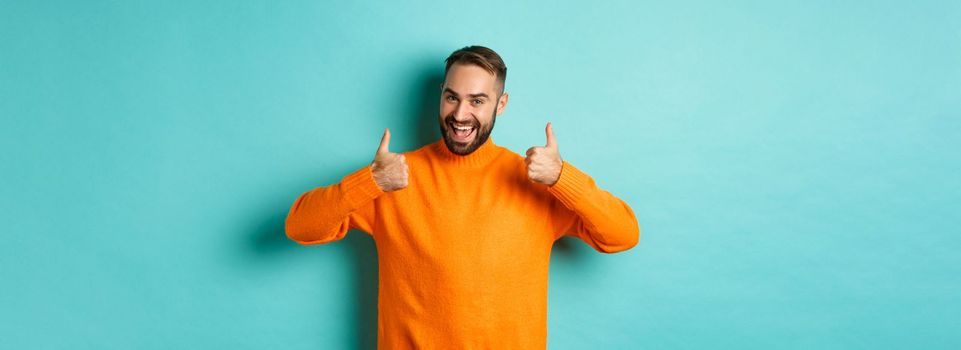 Handsome man expressing support, showing thumb up, encourage you, praise excellent work, approve and agree, standing over light blue background.