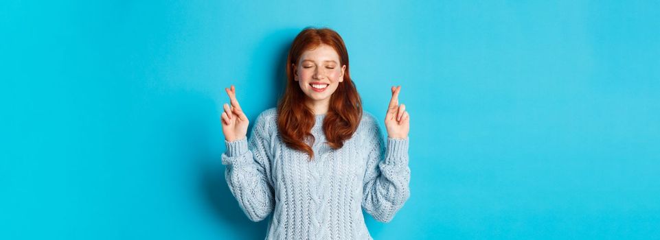 Hopeful redhead girl making a wish, cross fingers for good luck, smiling and anticipating good news or positive result, standing against blue background.