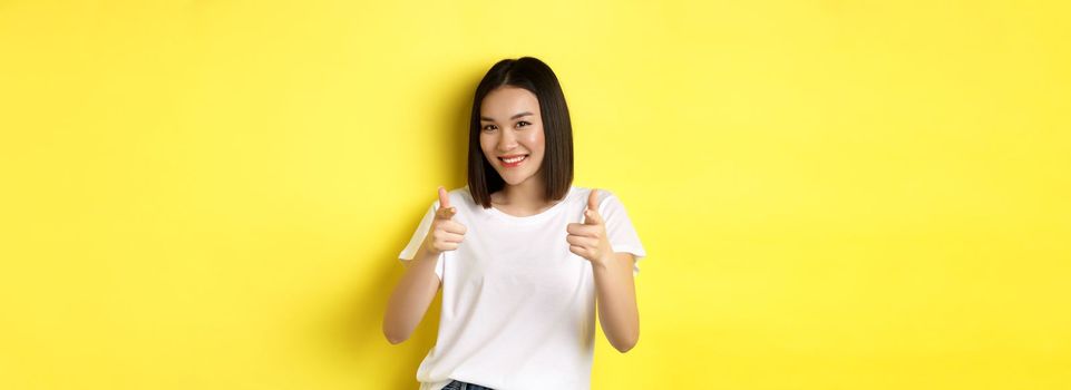 Pretty young asian woman in white t-shirt, pointing fingers at camera and smiling, praising or choosing you, say congrats, standing over yellow background.