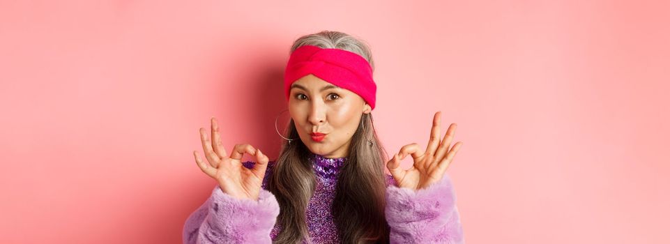 Fashion. Coquettish asian senior woman in trendy headband and faux fur coat, showing okay signs and pucker lips in kiss, praising something perfect, pink background.