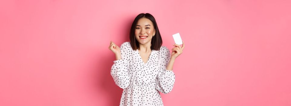 Shopping concept. Pretty asian woman holding bank credit card and smiling, standing over pink background.