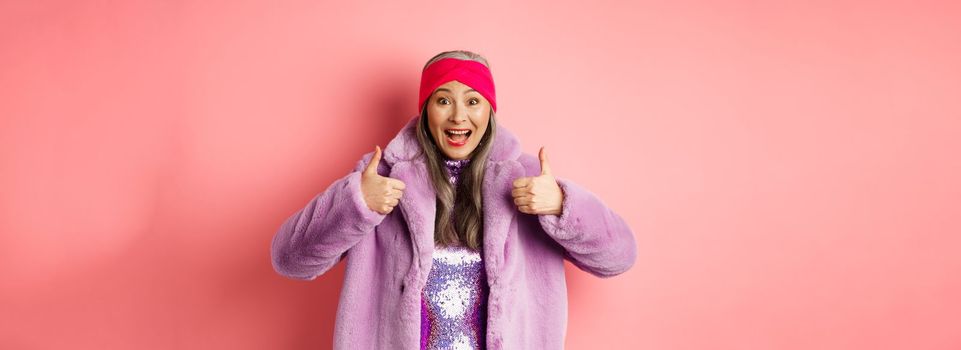 Fashion and shopping concept. Cheerful asian old woman showing thumbs-up in approval, like and recommend promotion, standing in stylish purple fur coat, pink background.