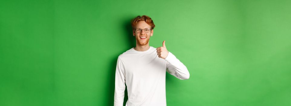 Handsome young man in glasses with red hair, showing thumbs-up and smiling satisfied, praising good eyewear store, standing over green background.