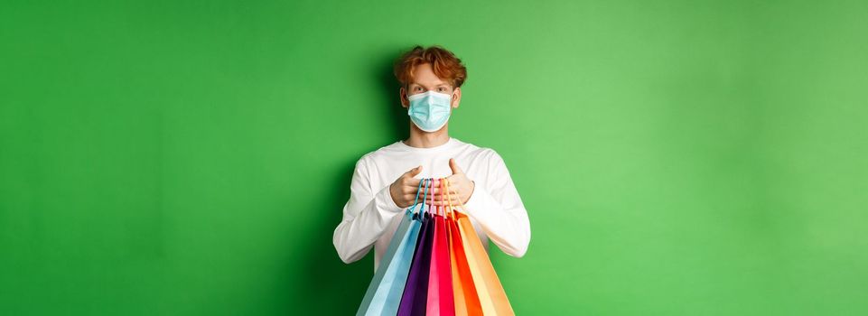 Pandemic and lifestyle concept. Cheerful redhead man going shopping in store, wearing medical mask and holding bags, standing over green background.