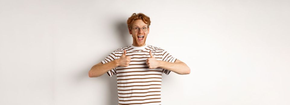 Young amazed man in red hair checking out something awesome, saying yes and showing thumbs-up, standing over white background.