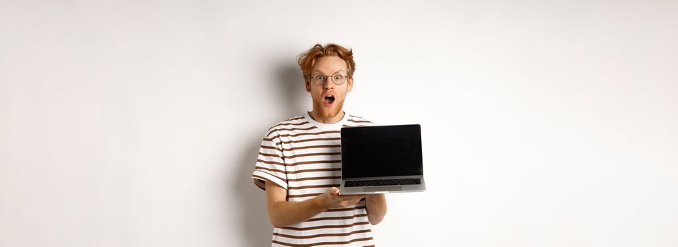 Excited redhead man drop jaw, showing laptop screen logo or website, demonstrate promo offer on display, standing against white background.