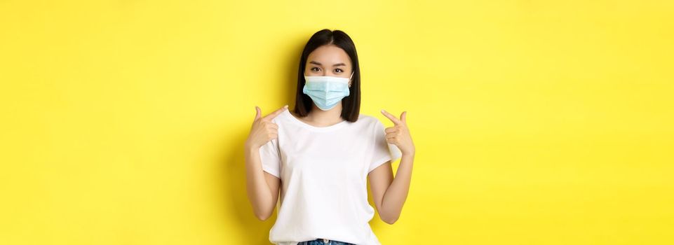 Covid-19, quarantine and social distancing concept. Young asian woman in white t-shirt, pointing at her medical mask from coronavirus, yellow background.
