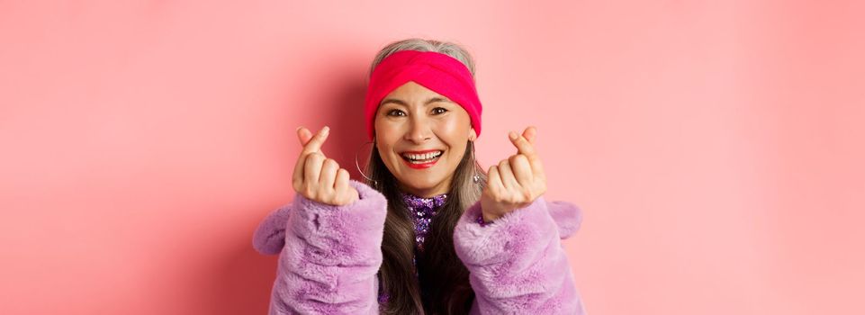 Fashion. Beautiful and fashionable asian senior woman smiling, showing korean heart gesture and looking happy, standing over pink background.