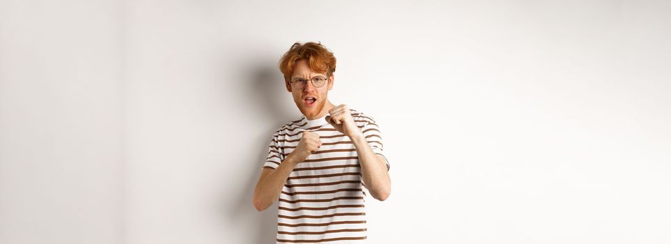 Funny young man with red hair raising fists for fight, shadow boxing and looking serious at camera, standing over white background.