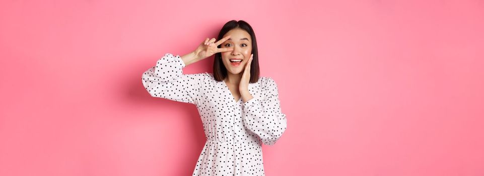 Beautiful and feminine asian woman showing kawaii sign on eye and looking amazed at camera, standing in dress over pink background.