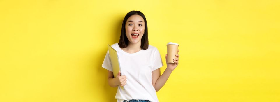 Happy asian woman drinking coffee and holding laptop, saying hi to you, standing over yellow background.