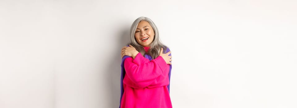 Valentines day and holidays concept. Lovely asian senior woman in pink sweater hugging herself with eyes closed, smiling, standing over white background.