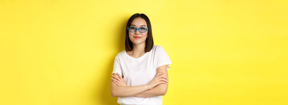Fashion and lifestyle concept. Sassy and confident asian woman in trendy sunglasses looking self-assured at camera, standing over yellow background.