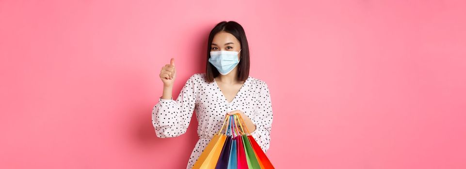 Covid-19, pandemic and lifestyle concept. Beautiful asian woman shopping in malls in medical mask, holding bags and showing thumb-up, standing over pink background.