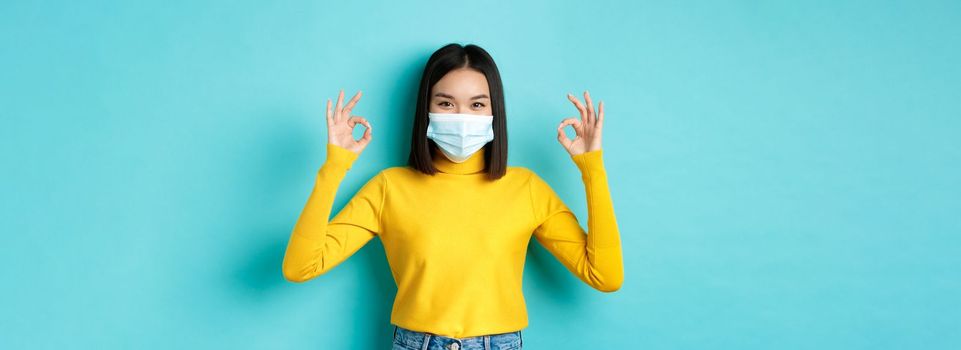 Covid-19, social distancing and pandemic concept. Smiling confident asian woman in medical mask showing OK signs, approve or praise good deal, standing over blue background.