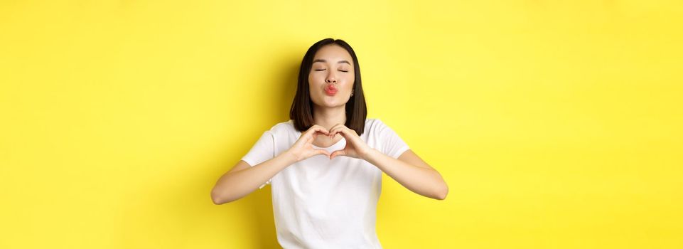 Beautiful asian woman showing I love you heart gesture, smiling at camera, standing against yellow background. Concept of valentines day and romance.