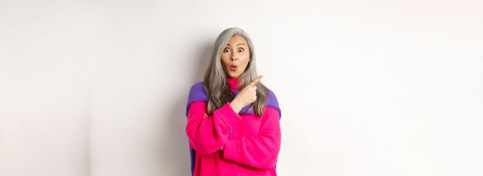 Portrait of impressed asian lady checking out promo offer, pointing at upper left corner and staring at camera amazed, standing in trendy pink sweater over white background.