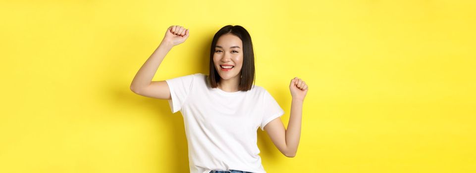 Carefree asian girl dancing and having fun, posing in white t-shirt against yellow background.