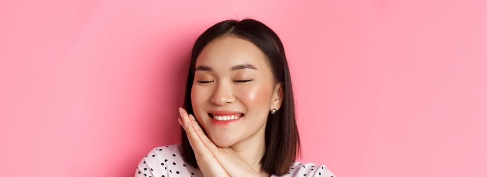 Beauty and skin care concept. Headshot of adorable and dreamy asian woman close eyes, smiling nostalgic, standing against pink background.