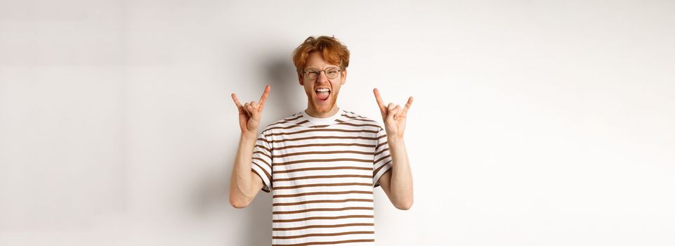 Funny and happy redhead man having fun, showing rock-n-roll horn and sticking tongue, enjoying party, standing over white background.