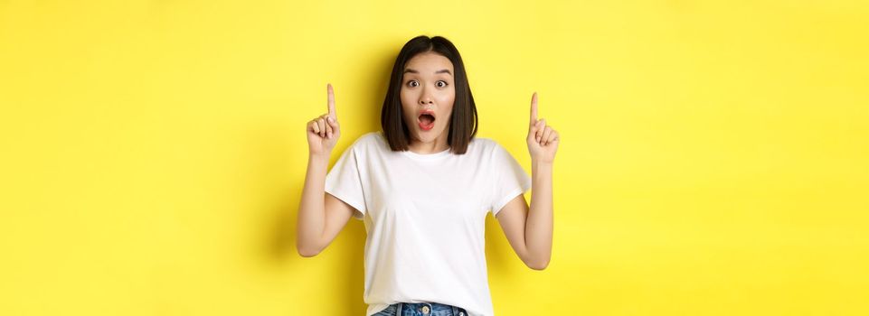 Beauty and fashion concept. Beautiful asian woman in white t-shirt pointing fingers up, standing over yellow background.