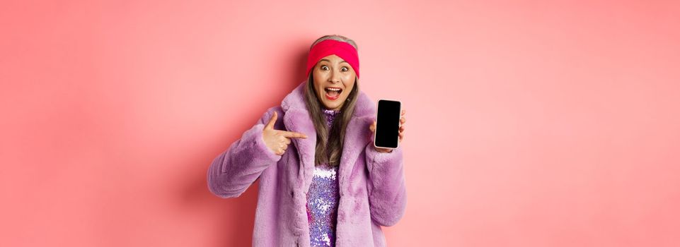 Online shopping and fashion concept. Stylish asian senior woman in faux fur coat pointing finger at blank smartphone screen, smiling happy, showing application on phone.