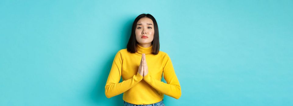 Young attractive asian girl begging for favour, need something and asking please, holding hands in pray, standing over blue background.