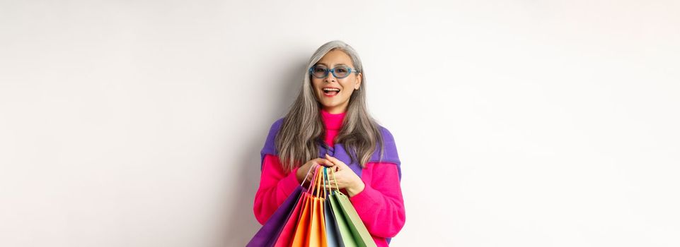Stylish senior asian woman in sunglasses going shopping on holiday sale, holding paper bags and smiling, standing over white background.