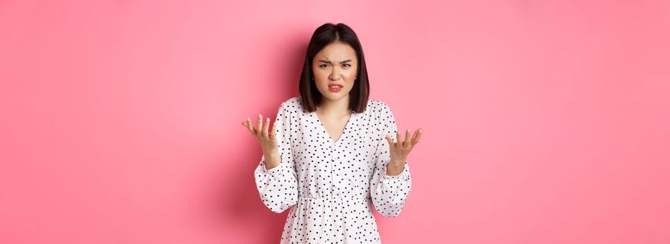 Angry and confused asian woman pointing hands at camera and grimacing furious, standing annoyed against pink background.
