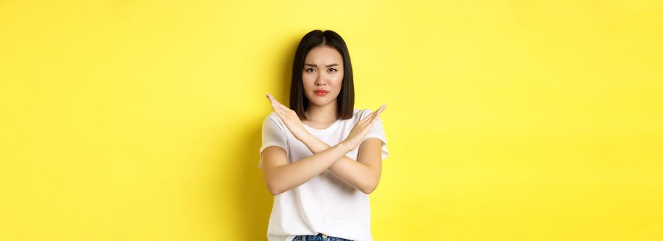 Serious and assertive korean woman showing cross stop gesture, frowning and telling no, prohibit action, disapprove something bad, standing over yellow background.