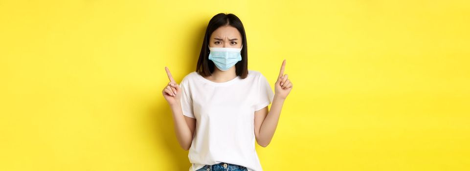 Covid-19, pandemic and social distancing concept. Disappointed asian girl in medical mask, frowning upset and pointing fingers up at logo, standing over yellow background.